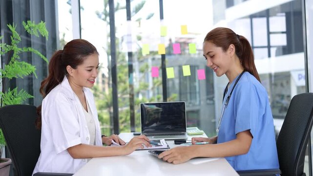 Asian female doctor and nurse discussing patient diagnosis with laptop and tablet in a modern hospital office.