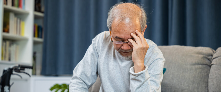 Asian mature old man sitting on sofa headache at home in living room, Depressed senior man unhappy hands rubbing eye suffering from a migraine