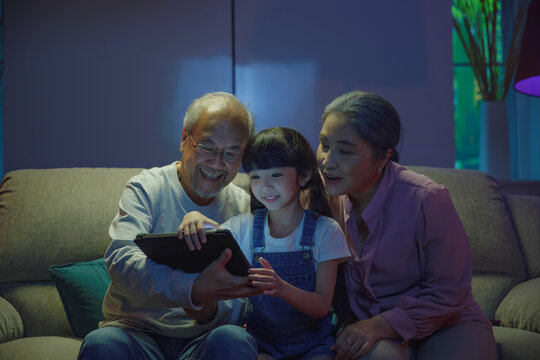 Happy Asian family looking video on digital tablet on a cozy couch at night, grandmother and grandfather playing with they little child girl on PC tablet on sofa