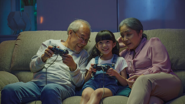 Happy Asian family bonding over a video game on a cozy couch, little girl playing video games console with senior grandparents together with controller joysticks