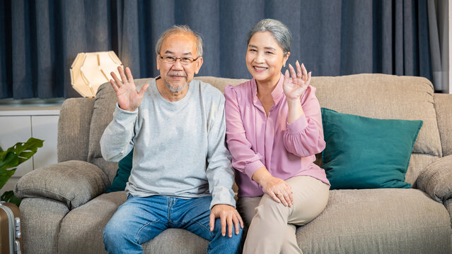 Happy Asian senior couple greeting someone while sitting on a comfortable couch, old man and woman sit close together on a sofa