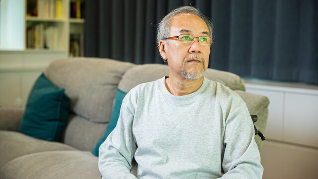 Asian senior man disabled sitting alone on wheelchair in living room looking out window at home and thinking, lonely elder sad old man retirement memory loss or nostalgia thought