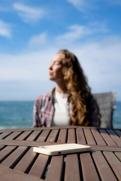 Smartphone in focus on a seaside table with a relaxed woman in the background, illustrating digital detox, unplugging and mindful living.