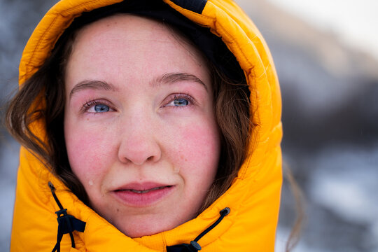 Emotional close-up portrait of a female mountaineer in a yellow down jacket looking into the distance during a winter mountain expedition.