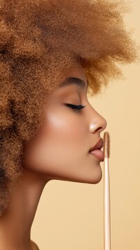 Profile of woman with beautiful coily hair and glowing skin. She holds a cosmetic brush near her lips, eye closed, on a beige background.
