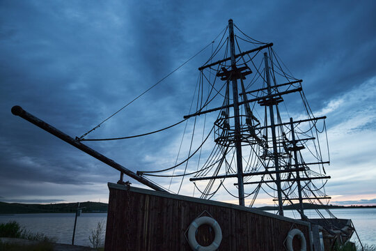 Blue-toned twilight lakeside landscape with antique-style wooden two-masted sailboats moored on the lake.