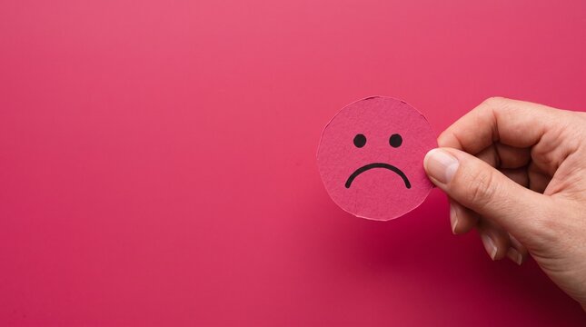 A person holds a small circular piece of pink paper with a hand-drawn sad frown face against a solid vibrant pink background representing negative mood