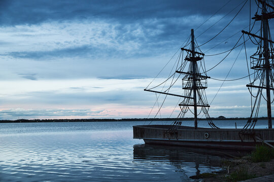 Blue-toned twilight lakeside landscape with antique-style wooden two-masted sailboats moored on the lake.