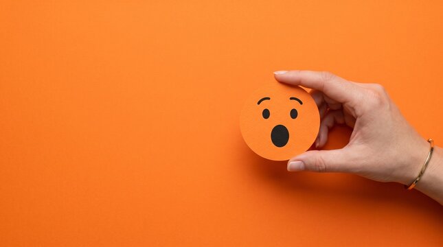 A Person Holding an Orange Paper Cutout Depicting a Surprised Emoji Face Against a Vibrant Solid Colored Background Space