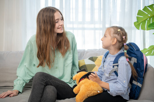 Young mother talking with her little daughter wearing backpack on sofa before school. Loving mother chatting with cute schoolgirl holding teddy bear in living room while preparing for school day..
