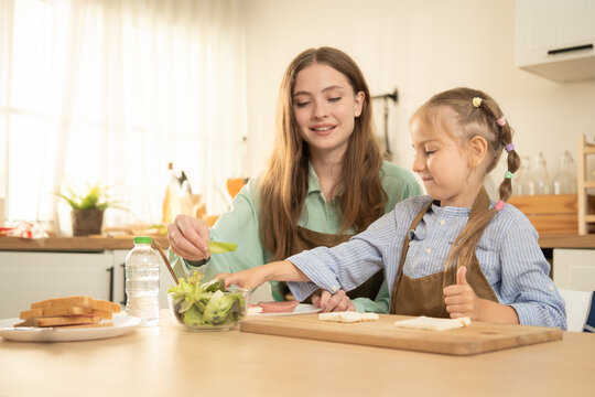Little girl giving thumbs up gesture while making healthy sandwich with mother in kitchen. Happy mother smiling at cute daughter showing thumbs up during fun cooking activity at wooden table.