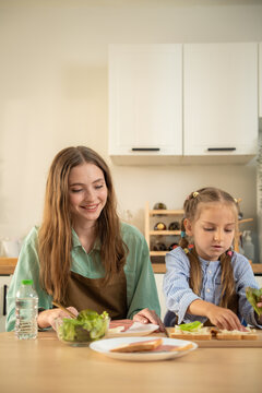 Mother and little daughter preparing healthy sandwich together at kitchen table. Cute young girl placing fresh lettuce on bread while mother supports her making healthy breakfast in home kitchen.