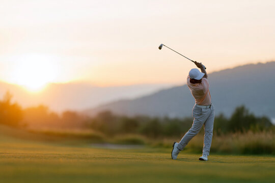 Golfer swings at golden sunset on serene golf course. Sun dips behind distant mountains, casting warm glow. Player wears light shirt, white cap, and gray trousers
