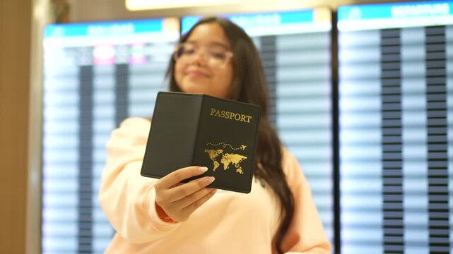 A young hispanic woman stands inside an airport terminal while holding a passport toward the camera, with flight information boards blurred in the background.