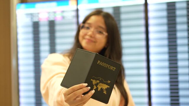 A young woman stands inside an airport terminal while holding a passport toward the camera, with flight information boards blurred in the background.