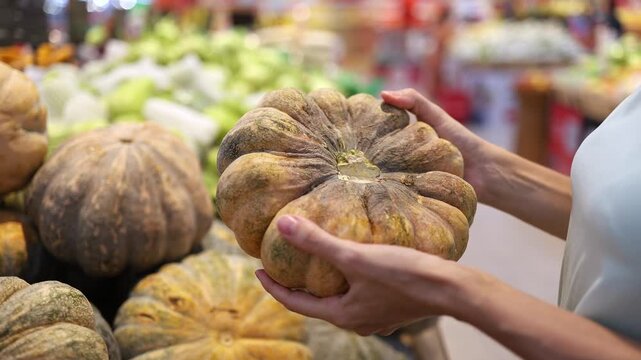 Close-up of a person holding and choosing a fresh ripe pumpkin or squash from a display at a supermarket or grocery store. Retail trade, food shopping and healthy nutrition concept.