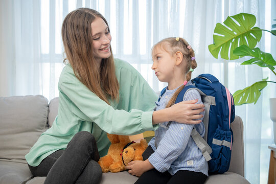 Caring mother holding her daughter's shoulder comforting her on living room sofa. Loving young mom encouraging her cute schoolgirl daughter wearing blue backpack before sending her to school.