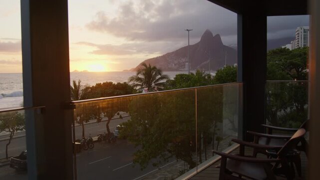 Golden sunset over Ipanema framed by the concrete columns and glass rail of a shaded terrace, a wooden chair angled towards the sea and the Dois Irmaos peaks.