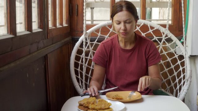 A calm woman thoughtfully eats a delicious homemade meal while sitting in a cozy hanging chair on a wooden balcony. She feels peaceful and relaxed enjoying her tasty dinner.