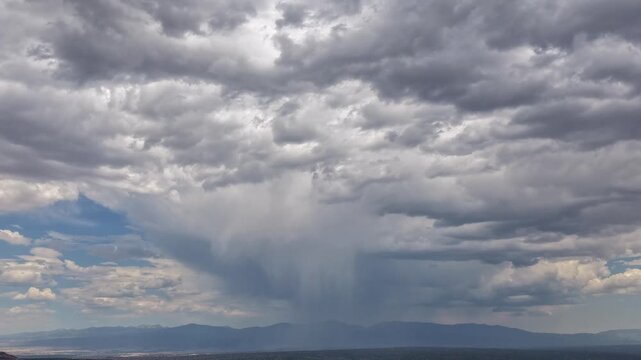 4K aerial timelapse capturing a heavy monsoon downpour sweeping across the high desert. A massive curtain of rain falls from dark, turbulent storm clouds toward the distant Southern Rocky Mountains.