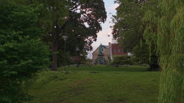 Drone footage unveiling the Hans Christian &Oslash;rsted monument through the greenery of Copenhagen's central park, with scenic pathways, trees, and city buildings beyond.