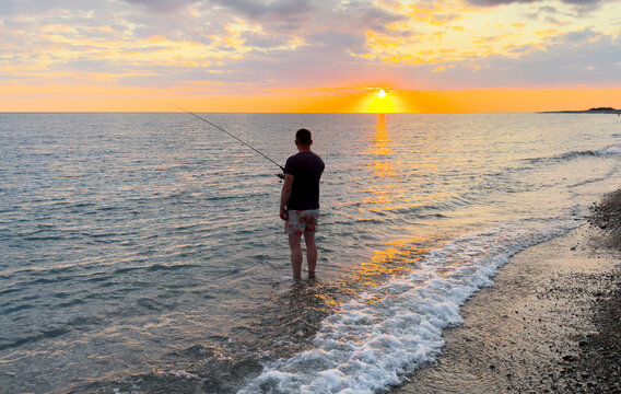 Fisherman on the beach with fishing rod standing on sea water.  Fishing at sunset with beautiful orange sky.