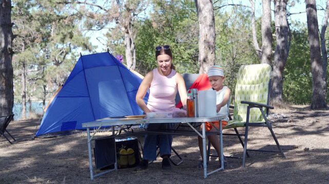 A happy mother and her joyful son prepare a picnic at a folding table near a blue tent. They feel content bonding during their camping trip. The sunny pine forest campsite feels peaceful.