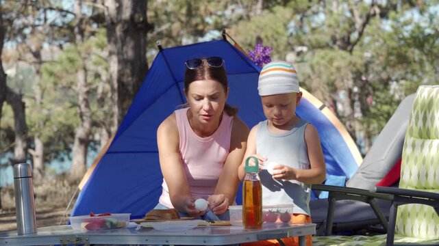 A caring mother and son joyfully prepare a picnic meal at a sunny forest campsite next to a blue tent. They feel happy and content sharing a peaceful morning together.