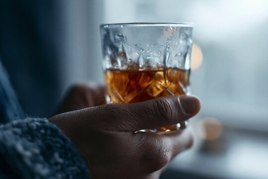Hands cradle a textured glass filled with amber liquid. Fingers grip the glass with quiet intensity. Light filters softly through a blurred window. Warm tones contrast with cool shadows around