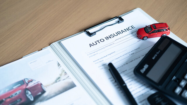Close-up of auto insurance document with red model car, calculator and pen on wooden desk, representing vehicle coverage and premium costs.