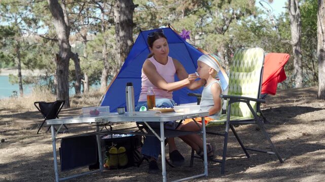 A loving mother gently wipes her son's face with a napkin at a picnic table. They feel content and happy during their lunch near a blue tent. The sunny pine forest campsite feels peaceful.