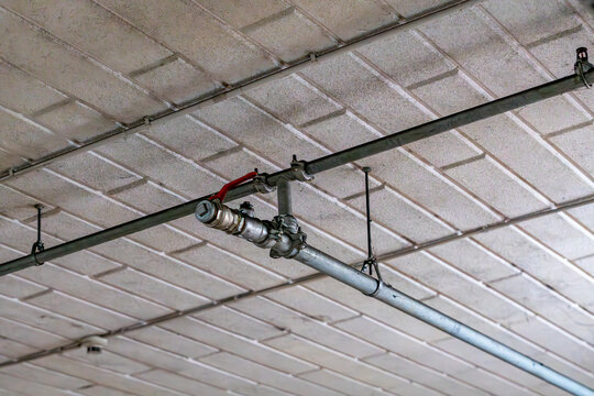 Metal pipe of water supply or fire sprinkler system with brass ball valve and red handle, mounted under concrete ceiling with steel clamps and threaded rods indoors.