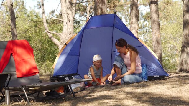 A loving mother and her son happily rest and chat inside a blue tent at a sunny forest campsite. They feel peaceful and content sharing a cozy morning together.