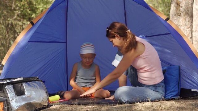 A loving mother and her playful son share a funny moment while sitting inside a blue tent. They feel joyful and amused playing together at a sunny forest campsite.