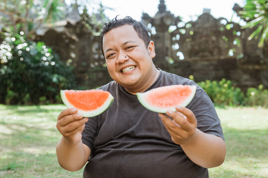 fat man smiles at the camera while holding watermelon slices in the outdoor background