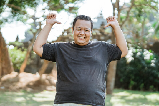Asian fat man standing with a self-pointing gesture with both thumbs in the outdoor background
