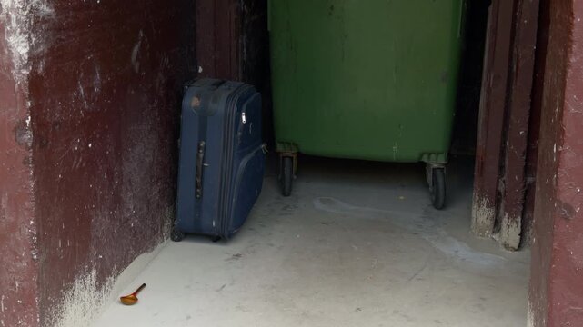 Blue suitcase beside green rolling bin inside narrow storage space with worn red walls