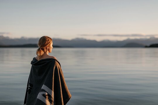 Woman wrapped in blanket looking at a calm lake at sunrise