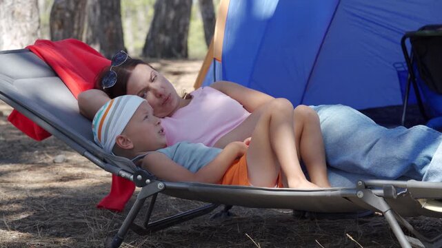 A mother and her young son rest together on a lounge chair while he points at something away. They share a tender, cozy moment in a shaded campsite in the woods.