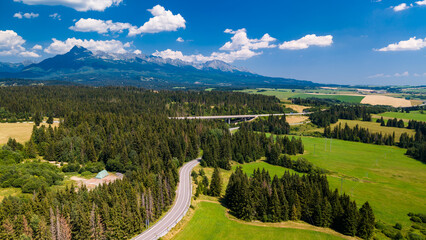 Fototapeta premium Mountains and green fields near a winding road in a rural area. A winding road runs through green fields and trees with mountains in the background.