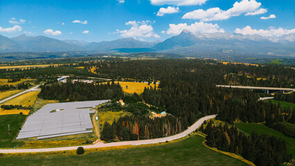 Fototapeta premium Solar panels sit beside a highway in a rural area surrounded by mountains. Solar panels cover land near a highway, with mountains in the background and fields around.