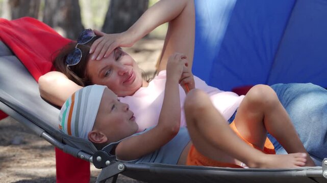 A happy mother and her young son lie together on a cot at a campsite. She smiles warmly as they share a sweet moment. They feel joyful, relaxed, and full of love.