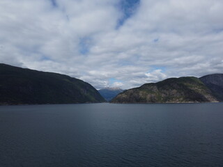 Majestic landscape view of a deep blue Norwegian fjord surrounded by dark forested mountains under a dramatic cloudy sky during daytime for travel and nature themes. © nikjuzaili