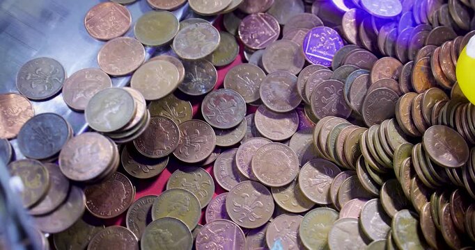 Slow motion footage showing a large collection of British pound coins inside an amusement arcade coin pusher machine with two pence pieces stacked and scattered beneath colourful illuminated lights
