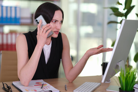 Stylish businesswoman in neat black suit talks on phone at desk. Successful office manager points at computer screen with confused expression