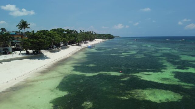 Drone pulls back and ascends to reveal Alona Beach, its white sand, shallow reef waters, palm-lined coast, and boats in Panglao, Bohol