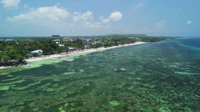 High-angle drone approaches Alona Beach, showing shallow reef waters, boats, white sand, and the Panglao coastline in Bohol