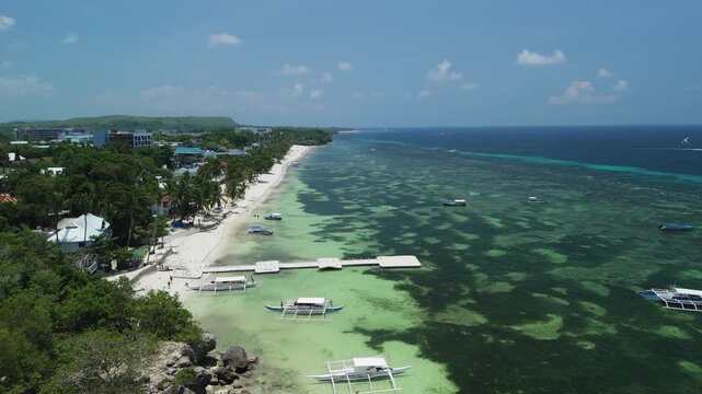 High-angle drone flyover shows a boat arriving at Alona Beach, with shallow reef water, white sand, and coastline in Panglao, Bohol