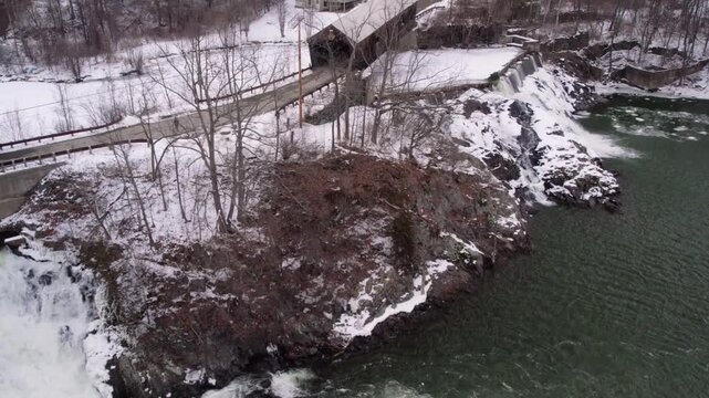 Aerial drone slowly orbiting from one historic Willard covered bridge toward the second bridge and waterfall along the snowy, partially frozen Ottauquechee River near White River Junction, Vermont.
