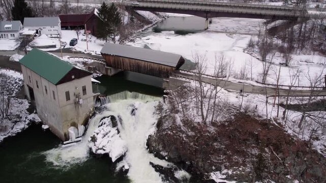Aerial drone shot slowly rotating left around a Willard covered bridge, waterfall and historic mill along the snowy, partially frozen Ottauquechee River near White River Junction, Vermont